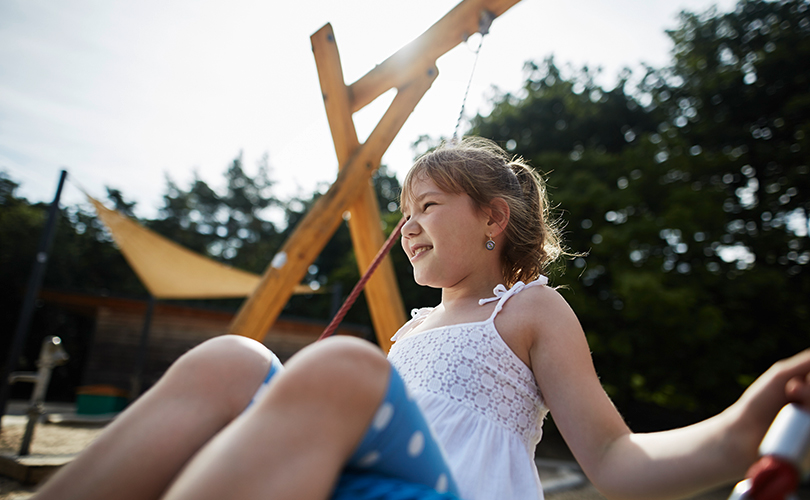Ein Mädchen schaukelt bei Sonnenschein auf einem Spielplatz.