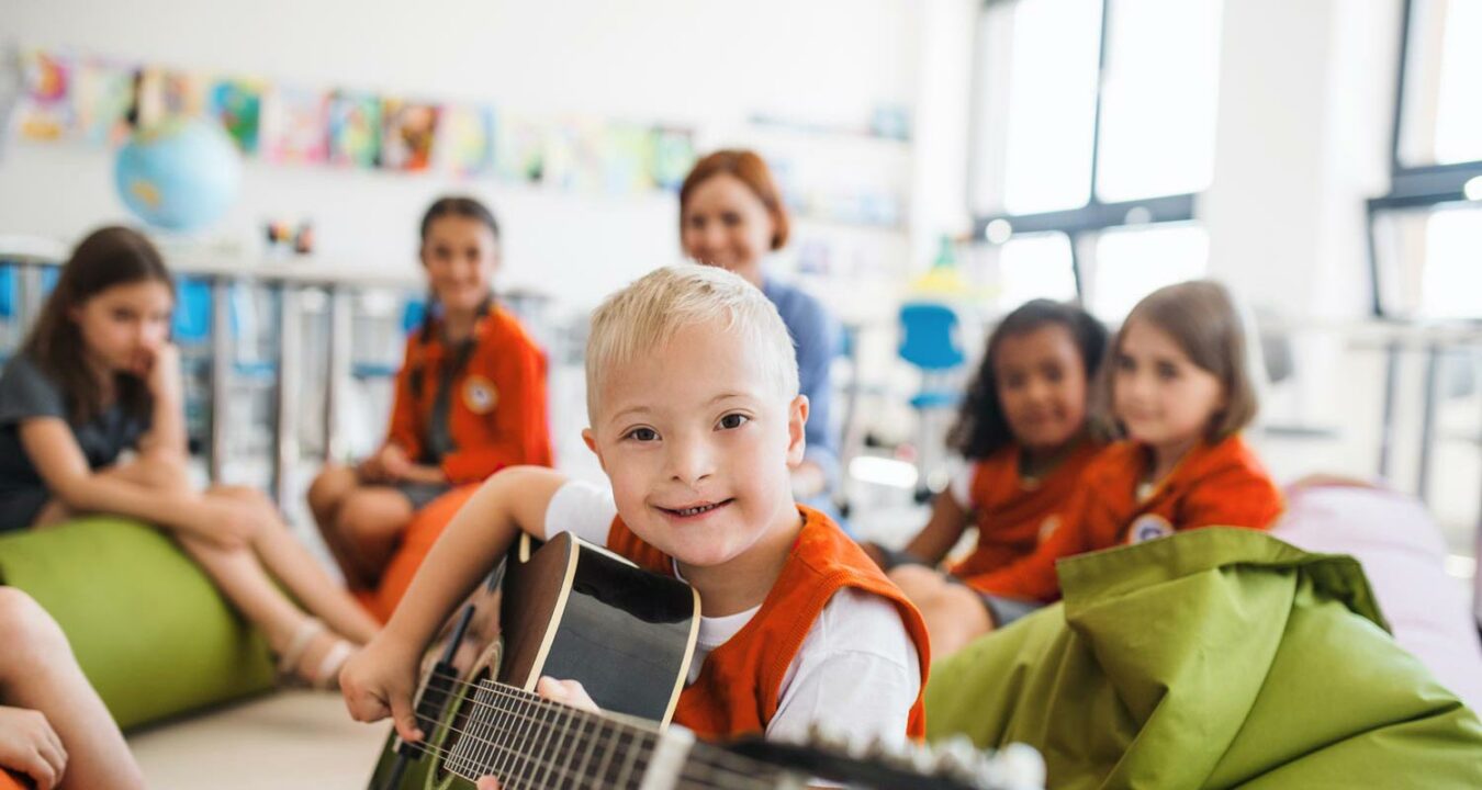 Das Bild zeigt eine bunte Gruppe Kinder und eine Betreuerin im Hintergrund. Ein kleiner Junge mit Downsyndrom blickt aufgeschlossen in die Kamera und spiel dabei Gitarre.