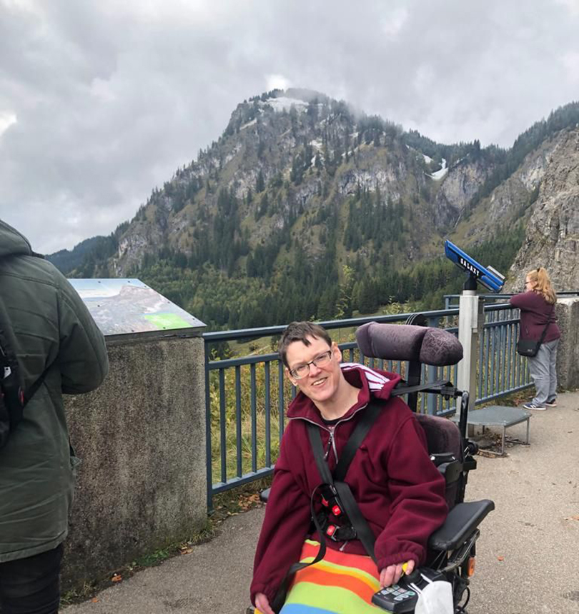 Eine Person im Rollstuhl blickt lachend in die Kamera. Im Hintergrund sieht man ein Bergpanorama.