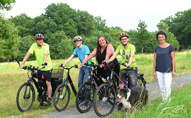 Eine Familie mit vier Personen macht eine Fahrradtour. Sie haben einen Hund dabei und eine Mitarbeiterin der Lebenshilfe.
