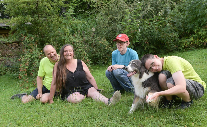 Eine Familie mit vier Personen sitzt auf der Wiese. Einer der Jungs kuschelt mit einem Hund.