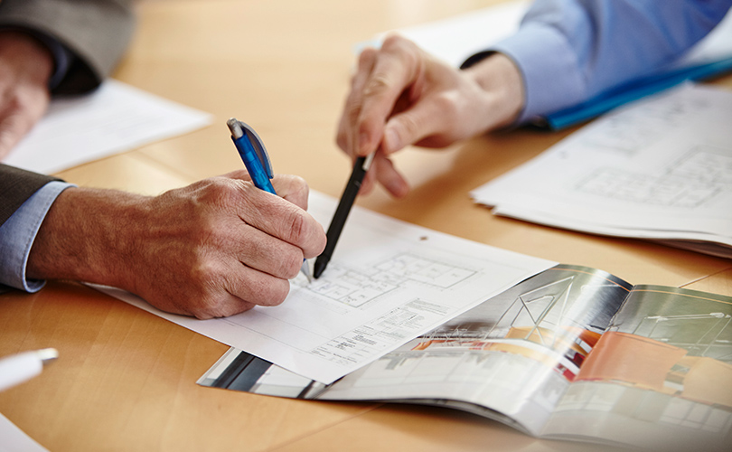 Zwei Personen zeigen mit Stiften auf ein Blatt Papier mit einer Grundrisszeichnung einer Wohnung.