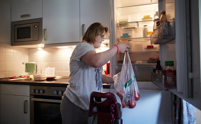 Eine Frau packt ihre Einkäuft in den geöffneten Kühlschrank. Sie hat einen Tüte mit Tomaten in der Hand.