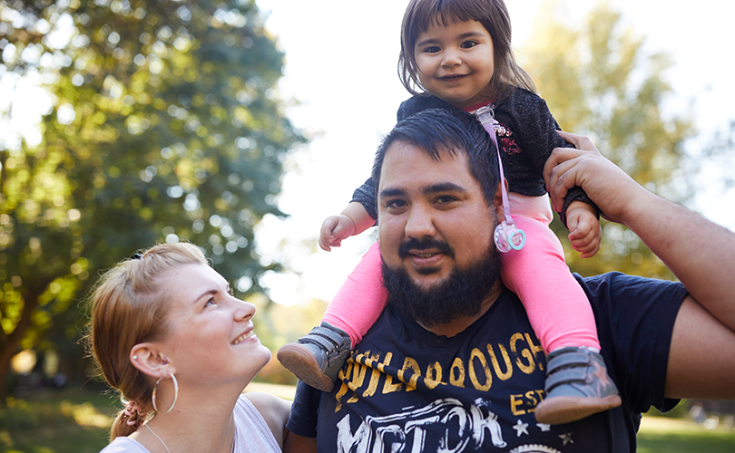 Eine Mutter, ein Vater und ein Kind sind im Park. Das Kind sitzt auf den Schultern des Vaters. Die Mutter schaut das Kind an.