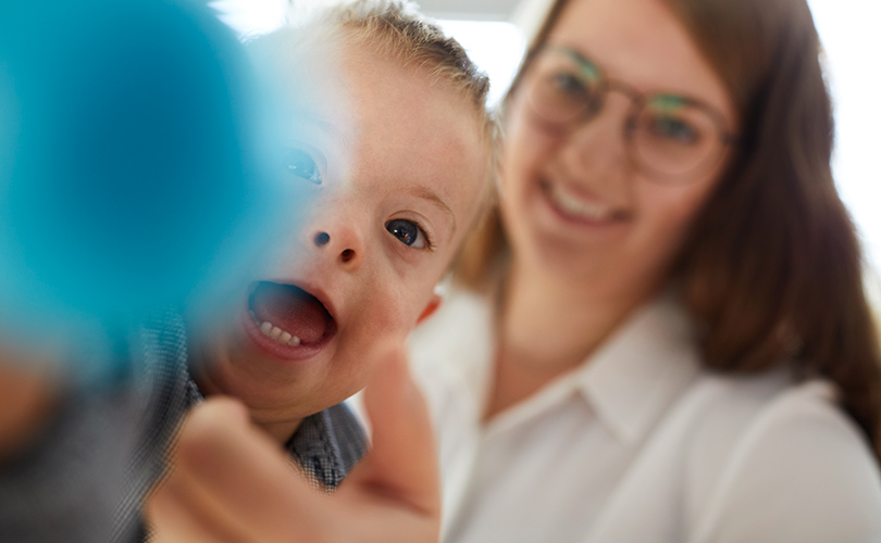 Eine Mutter und ihr Sohn spielen. Der Junge hält einen Ball in die Kamera. Die Mutter sitzt im Hintergrund und schaut zu.