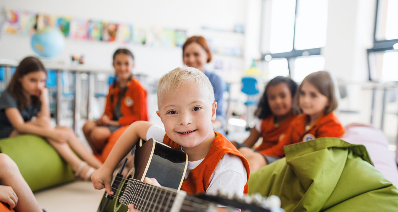 Das Bild zeigt eine bunte Gruppe Kinder und eine Betreuerin im Hintergrund. Ein kleiner Junge mit Downsyndrom blickt aufgeschlossen in die Kamera und spiel dabei Gitarre.