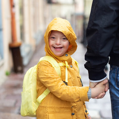 Ein Junge mit Trisomie 21 geht an der Hand eines Mannes im Regen spazieren. Er blickt fröhlich in die Kamera.