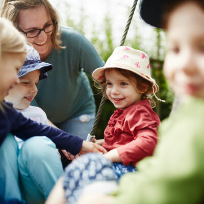 Mehrere Kinder spielen und schaukeln. Eine Betreuerin passt auf.