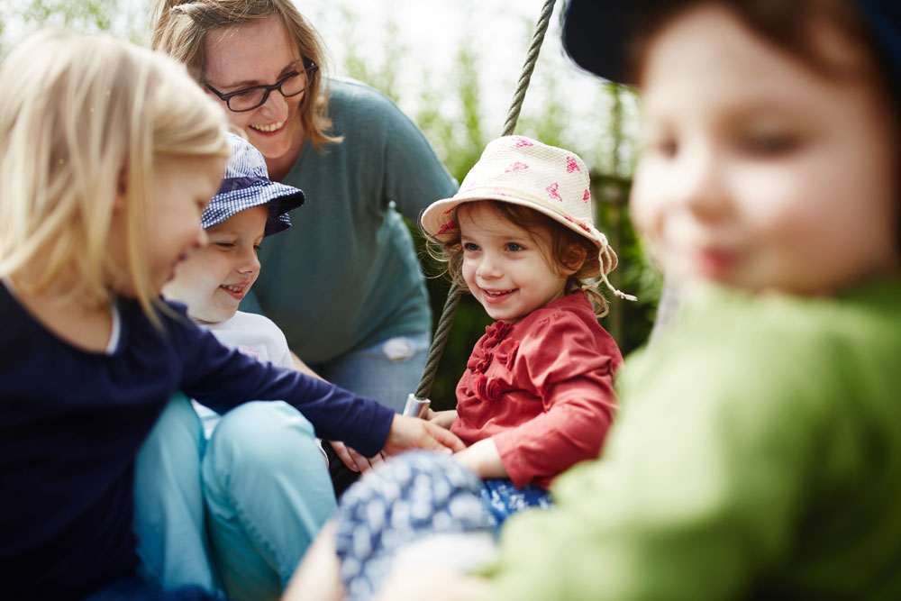 Mehrere Kinder spielen und schaukeln. Eine Betreuerin passt auf.