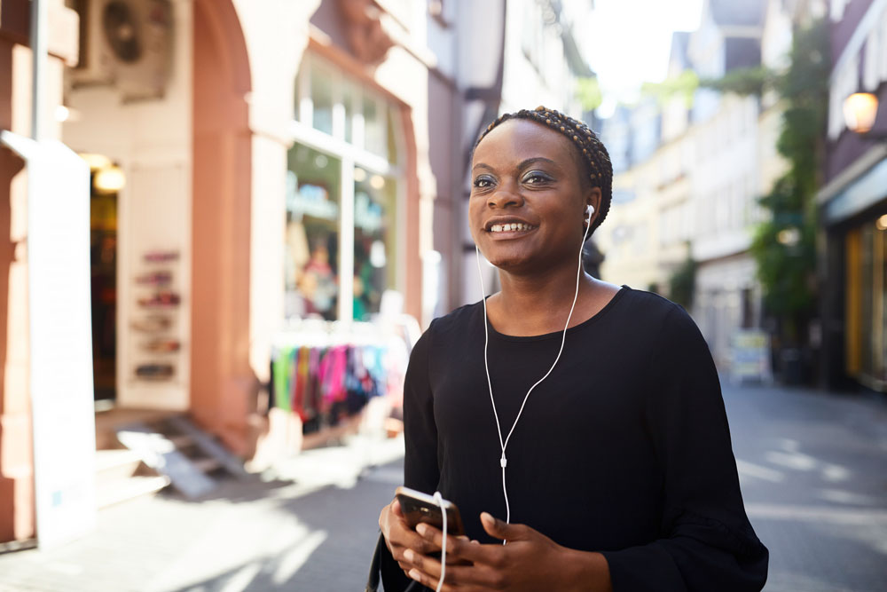 Eine junge Frau hat ein Handy in der Hand und Köpfhörer in den Ohren. Sie geht lächelnd durch die Fußgängerzone. 